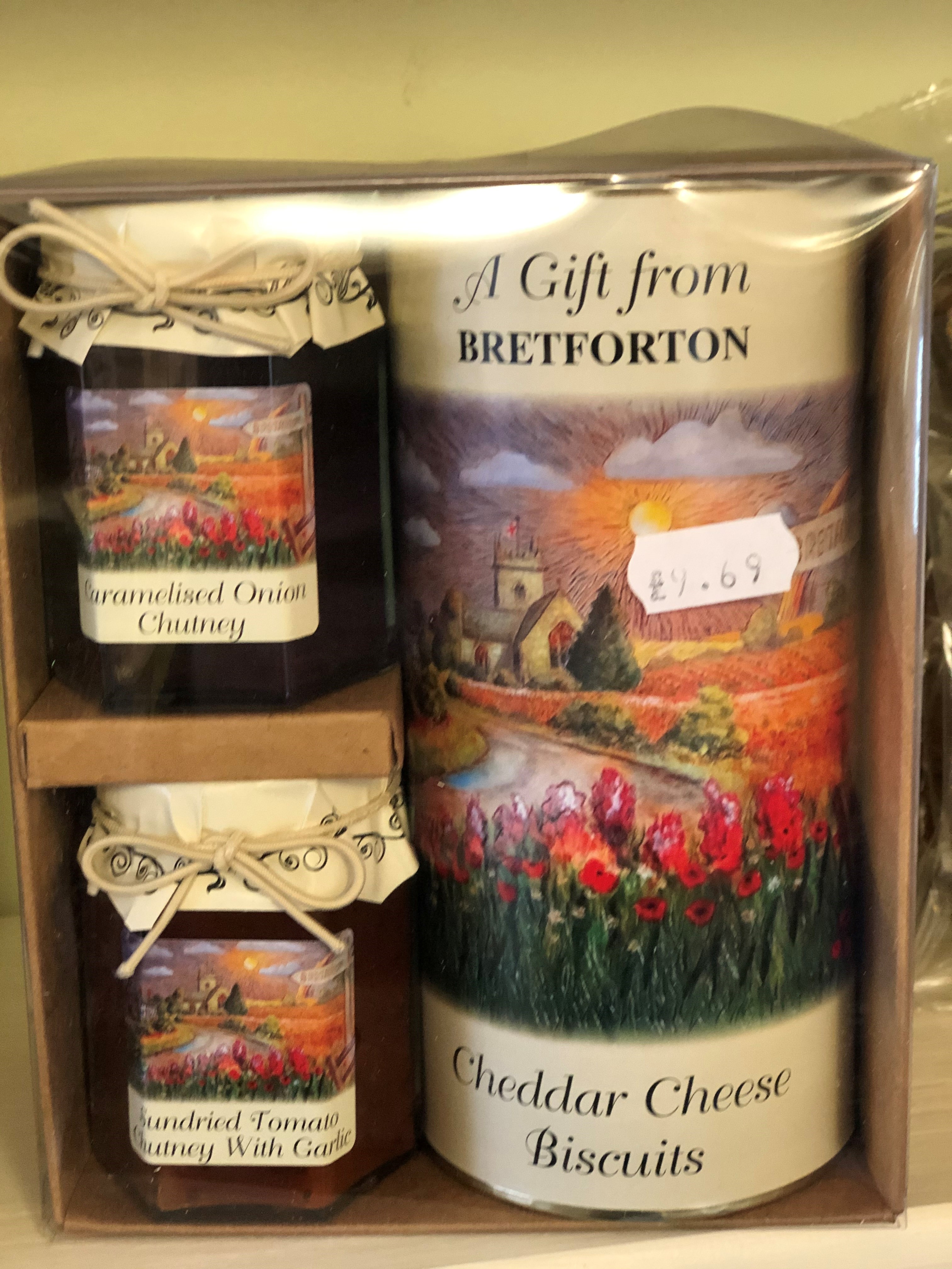 A photo of a gift pack of "Gifts from Bretforton", showing jars of caramelised onion chutney, sundried tomato chutney and a tube of cheddar cheese biscuits. Gifts have a reproduction of the oil painting of Bretforton that is displayed in the Shop on them