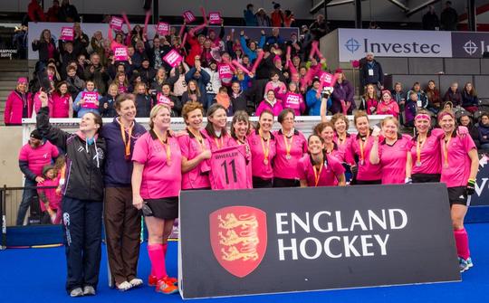 A photo of Bretforton ladies hockey team with their supporters in the background, posing behind an 'England Hockey' advertising board